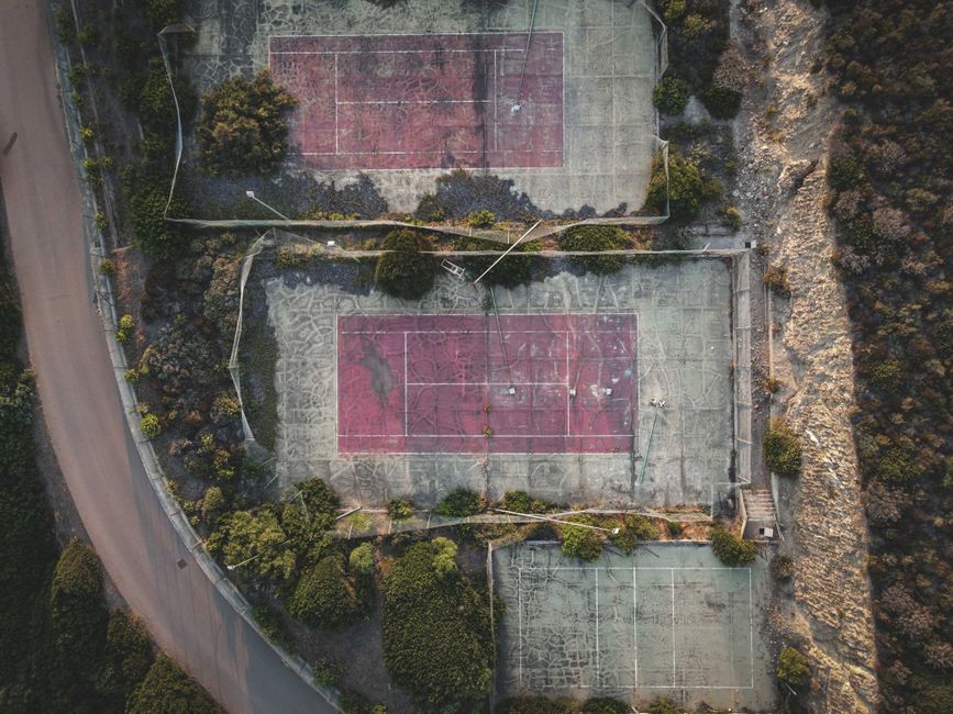 An aerial view of three outdoor basketball courts adjacent to a paved road, with the courts covered in faded red and green paint markings. The courts are surrounded by overgrown trees and bushes, creating a partially enclosed environment. One of the courts appears to have a damaged surface with patches of worn or missing paint, while the others are in slightly better condition. The courts are enclosed by a low chain-link fence, some of which is covered with netting or fencing material. The surrounding area features natural terrain and vegetation, indicating that the courts are situated in a park or recreational area. This scene can be associated with local community sports or recreational activities, relevant to logistics and equipment transport involved in house removals or relocation services, such as Man with Van Earls Court.