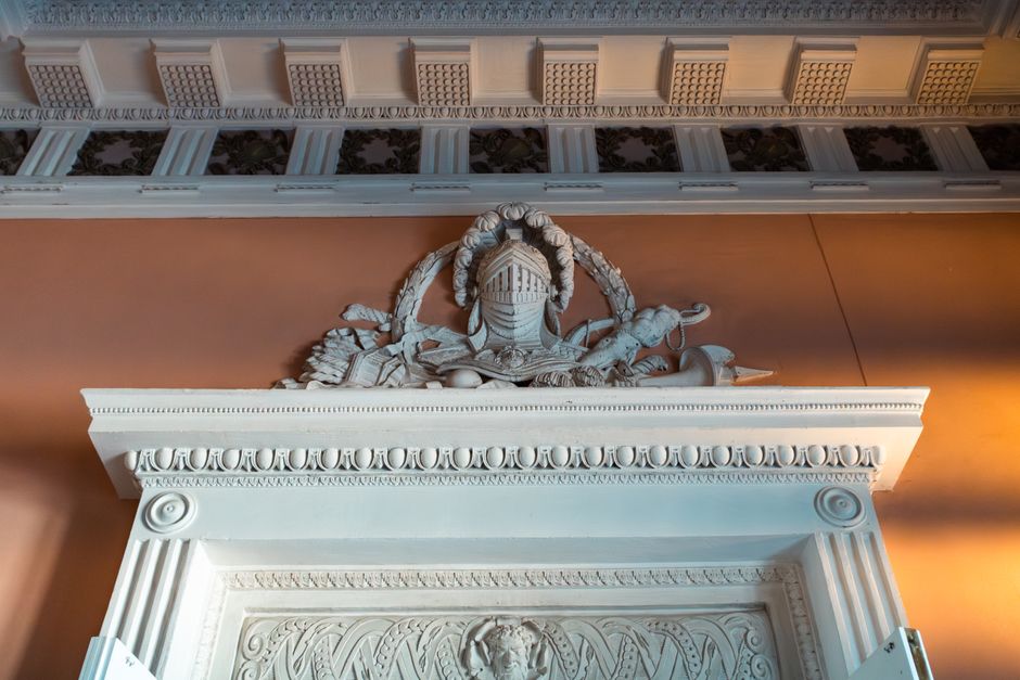 A close-up image of an ornately decorated interior fireplace mantel with classical architectural details, featuring intricate white sculptural embellishments on a pinkish-brown wall. Above the mantel, there is a detailed decorative frieze with a repeating pattern and a carved ornamental crest. The scene is lit with warm lighting, highlighting the craftsmanship of the cornice and molding. This interior detail, captured during a home relocation or furniture transport process, is part of a setting where items such as wooden furniture and packing materials might be displayed or prepared for moving. The image emphasizes the structural and decorative elements typical of period interiors, relevant to house removals and packing services provided by Man with Van Earls Court.
