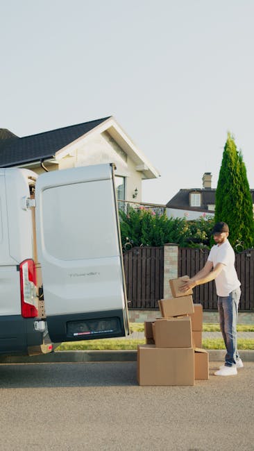 A man is standing on a residential street next to a white cargo van with its rear door open, preparing to load or unload cardboard boxes of various sizes. The boxes are placed on the pavement beside him, some stacked and others individually arranged. The man is carrying a small cardboard box, and a trolley is partially visible inside the van, indicating an ongoing packing or furniture transport process. The scene takes place during daylight hours, with a background of suburban houses, a wooden fence, and tall green shrubs or trees. The environment appears clean and organized, reflecting typical home relocation activities associated with professional removals services such as those offered by Man with Van Earls Court, supporting efficient packing and moving logistics.