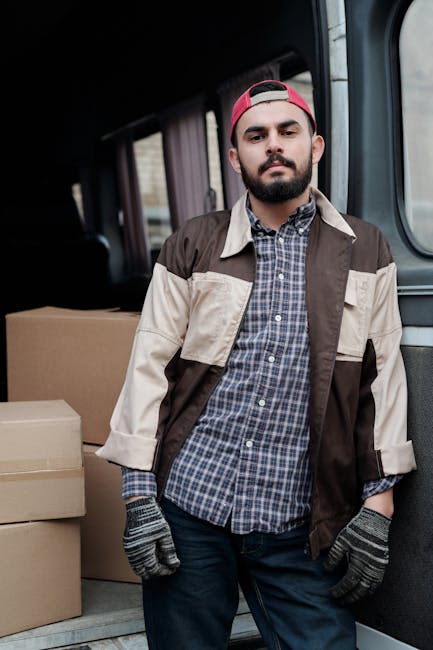 A man is standing on a residential street next to a white cargo van with its rear door open, preparing to load or unload cardboard boxes of various sizes. The boxes are placed on the pavement beside him, some stacked and others individually arranged. The man is carrying a small cardboard box, and a trolley is partially visible inside the van, indicating an ongoing packing or furniture transport process. The scene takes place during daylight hours, with a background of suburban houses, a wooden fence, and tall green shrubs or trees. The environment appears clean and organized, reflecting typical home relocation activities associated with professional removals services such as those offered by Man with Van Earls Court, supporting efficient packing and moving logistics.