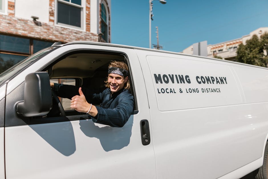 A young male mover with long hair, wearing a black bandana and a dark blue long-sleeve shirt, is sitting in the driver's seat of a white moving van with the label 'MOVING COMPANY LOCAL & LONG DISTANCE' on the side. He is smiling and giving a thumbs-up through the open window. The van is parked on a street, with a brick building and other modern structures visible in the background under a clear blue sky. Inside the vehicle, packing materials such as cardboard boxes and protective blankets are visible, suggesting an active home relocation or furniture transport process. The scene reflects professional packing and loading, with the mover ready to assist in house removals or moving logistics, as part of services provided by Man with Van Earls Court.