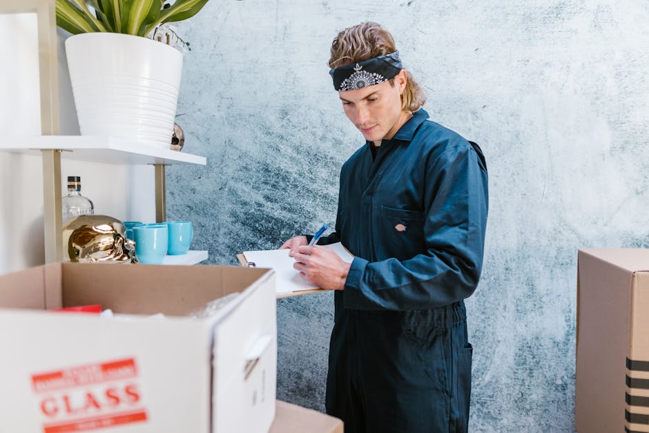 A man with a beard, wearing a black cap and a white shirt, standing inside a property next to a stack of cardboard boxes used for packing and moving. The boxes are arranged on a surface, some stacked higher than others, and are secured with brown packing tape. The man is holding a clipboard and pen, appearing to take notes or check inventory during a house removal or packing process. The environment suggests an indoor setting with natural lighting. This scene illustrates the packing and handling aspects of home relocation, as performed by Man with Van Earls Court, a professional removals service focused on furniture transport and moving logistics.