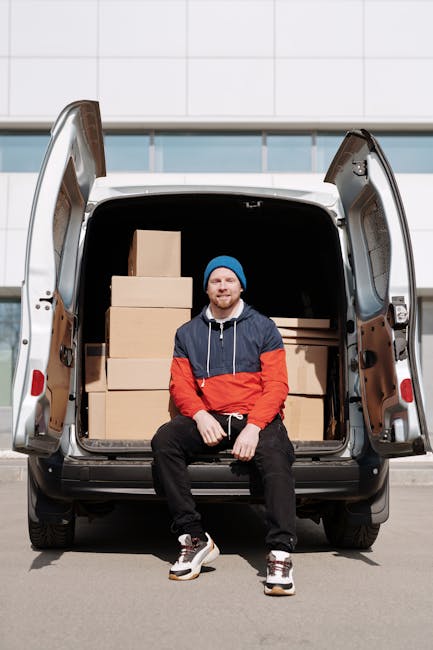 A man with a beard, dressed in a blue and red jacket, black pants, and a blue beanie, is sitting on the pavement in front of an open van loaded with neatly stacked cardboard boxes of various sizes. Th